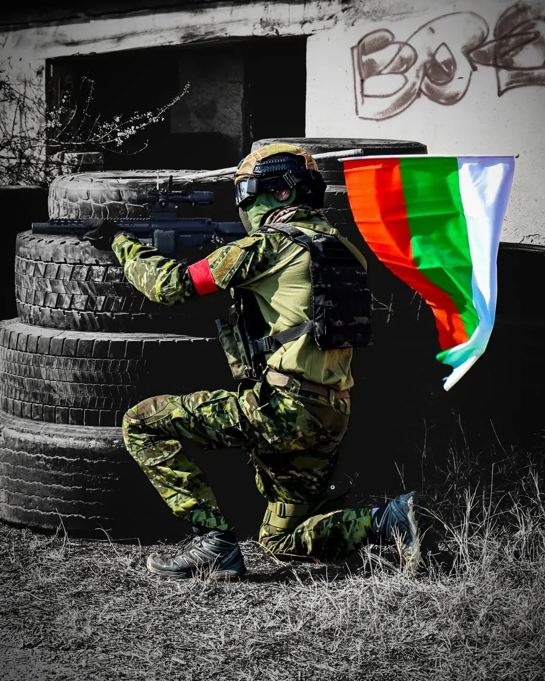 Niko from GFA Airsoft kneeling in cover with a Bulgarian flag during an airsoft game in Bulgaria